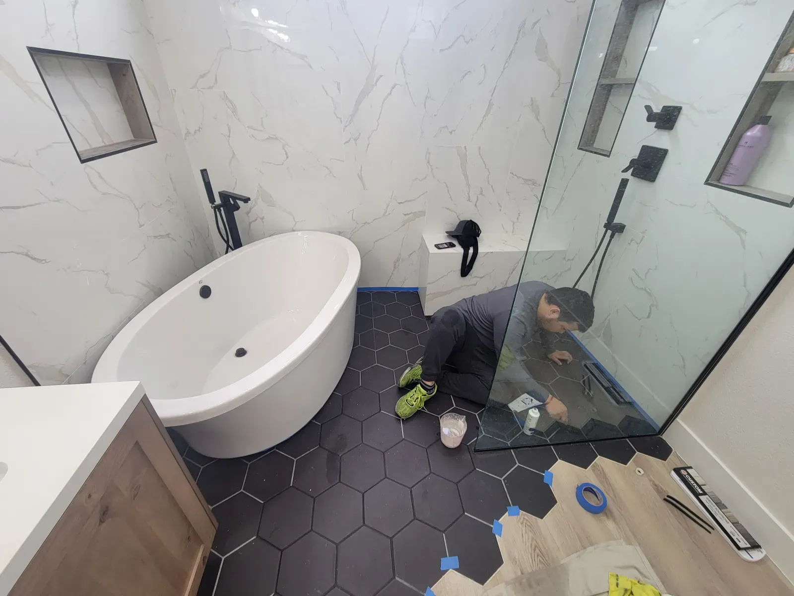 Technician sealing grout on a black hexagon tile bathroom floor beside a freestanding tub and glass shower